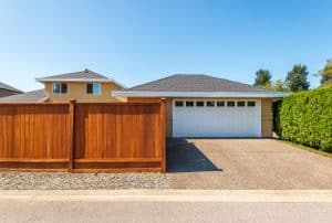 Single-story house with a white double garage door, wooden fence, and paved driveway, viewed from the street on a clear, sunny day.