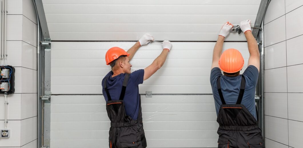 Two workers in orange hard hats and overalls installing a white sectional garage door panel.