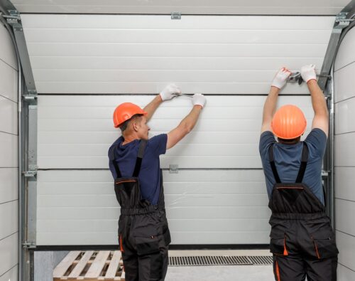 Two workers in orange hard hats and overalls installing a white sectional garage door panel.
