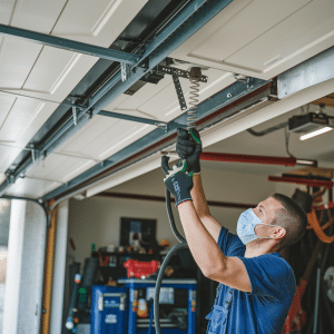 A technician wearing gloves and a face mask repairs the mechanism of a garage door inside a well-organized garage workspace.