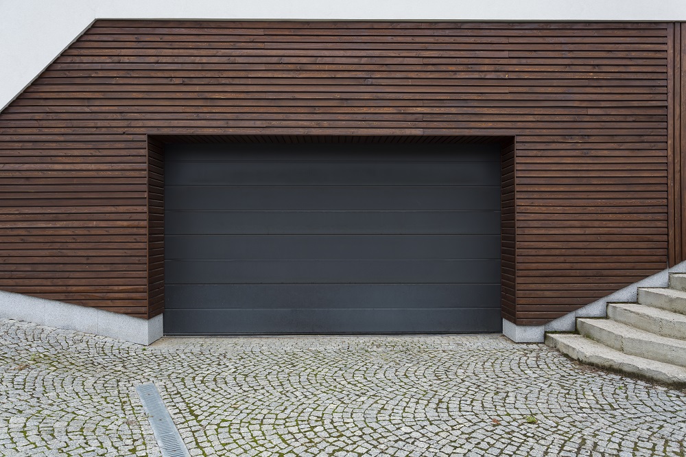 Modern garage with a dark gray door, surrounded by horizontal wooden paneling, adjacent to stone steps and a cobblestone driveway—perfect for those seeking top-notch garage door services in Parker.