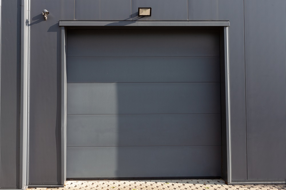 A closed gray industrial garage door set in a metal building, featuring Garage Door Wind-Load Reinforcement in Lakewood, with a security camera and light fixture mounted above.