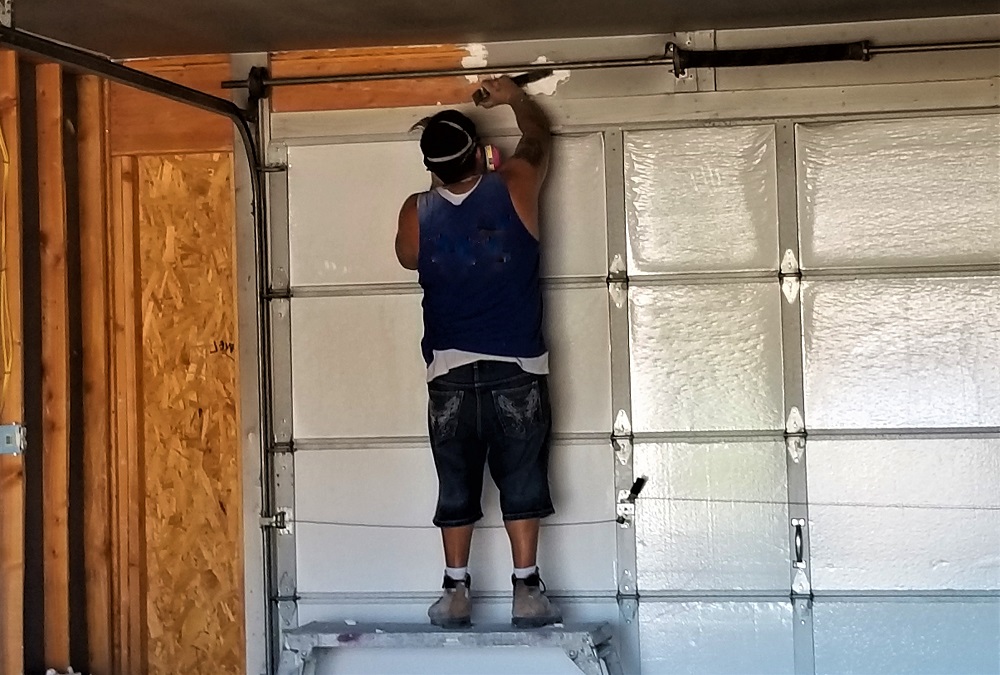 A person stands on a ladder, painting or repairing the upper part of a garage wall in a blue tank top, shorts, and safety gear—possibly preparing the space before installing the Best Insulation for Garage Doors.