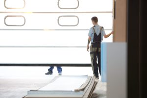 Two workers stand near a large, partially opened garage door with construction materials on the floor in front of them, appearing to discuss a garage door repair.