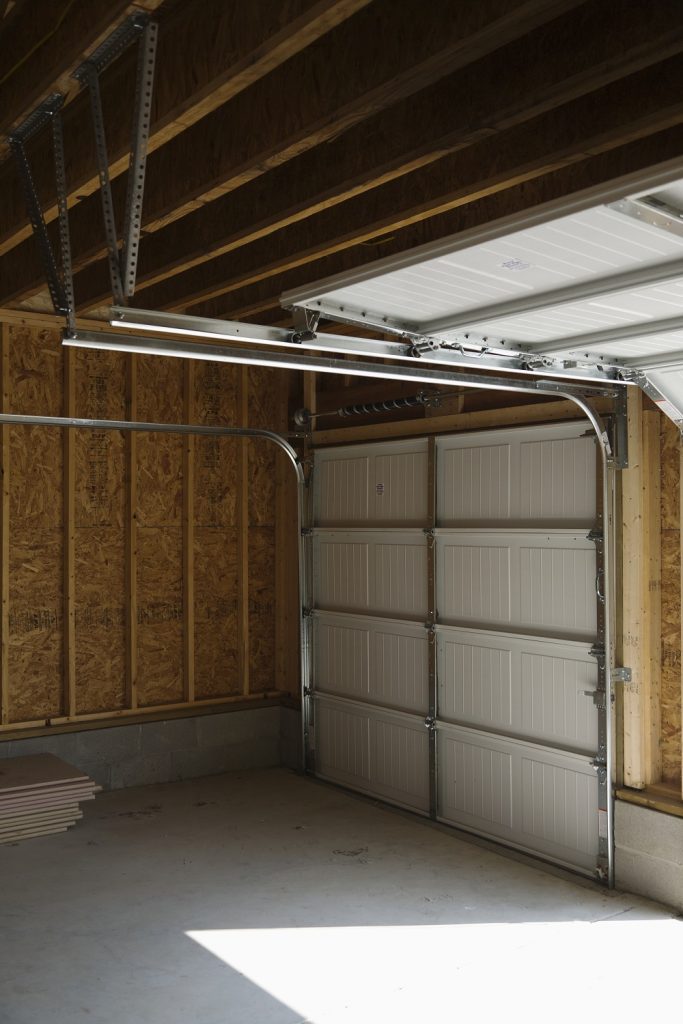 	
An empty garage with unfinished walls, an open ceiling exposing beams, and a closed sectional door with sunlight streaming in—ideal for adding Garage Door Wind-Load Reinforcement in Lakewood.
