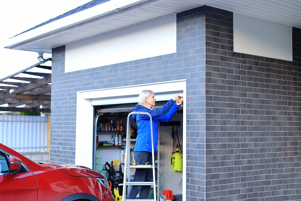 A person stands on a ladder, measuring the exterior of a brick garage near a red car, with tools and equipment visible inside—perhaps checking why garage doors stuck halfway open in Parker.