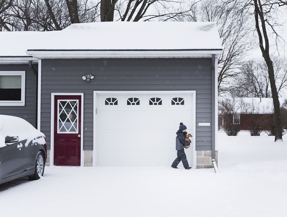A child in winter clothing walks a small dog on a snowy driveway in front of a gray house with a red door and a Garage Door in Parker.