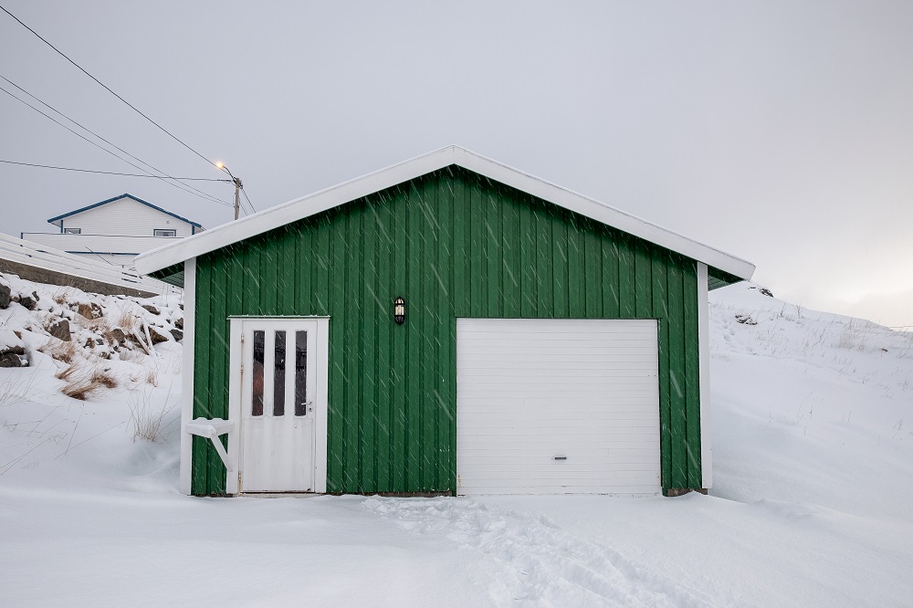 A small green shed with a white door and garage door in Parker stands in a snowy landscape, with footprints in the snow leading to the entrance.