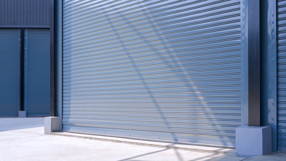 A large closed metallic roller shutter door on a modern industrial building, with sunlight casting shadows on the surface—built tough to withstand even harsh hailstorms in Parker.