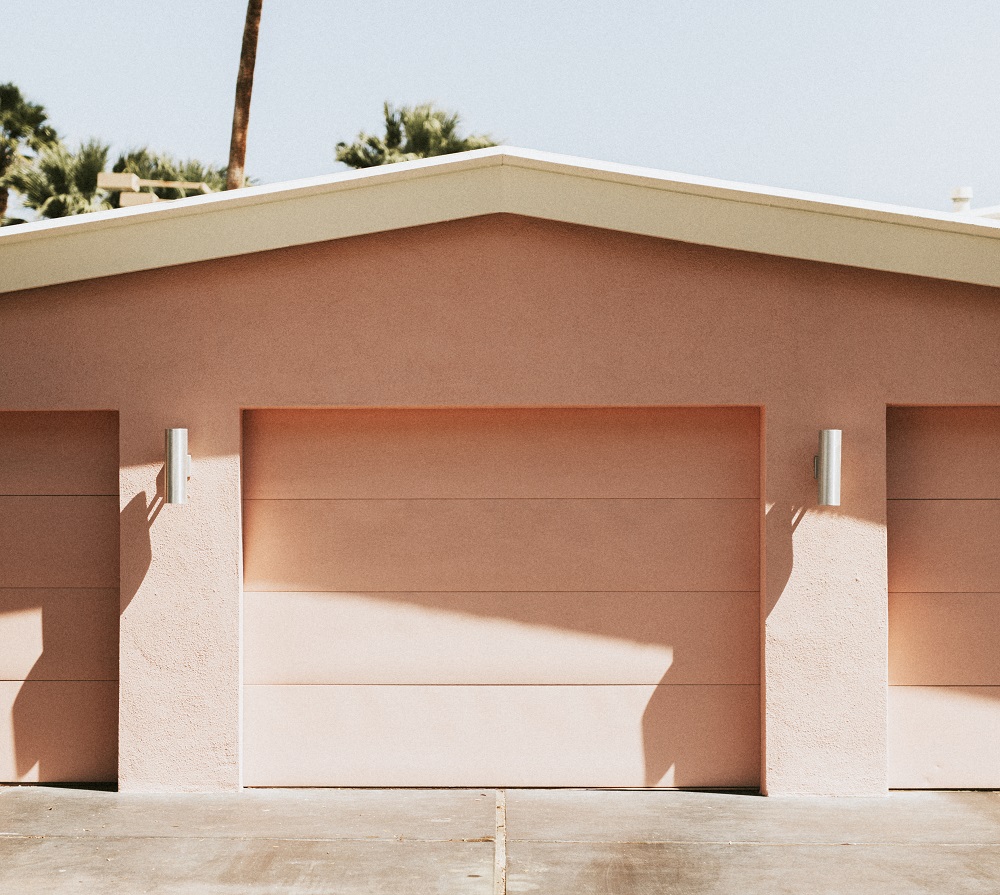 A pink garage with closed doors, white trim, and two silver wall-mounted lights stands ready for Same-Day Garage Door Service in Lakewood, with palm trees visible in the background.