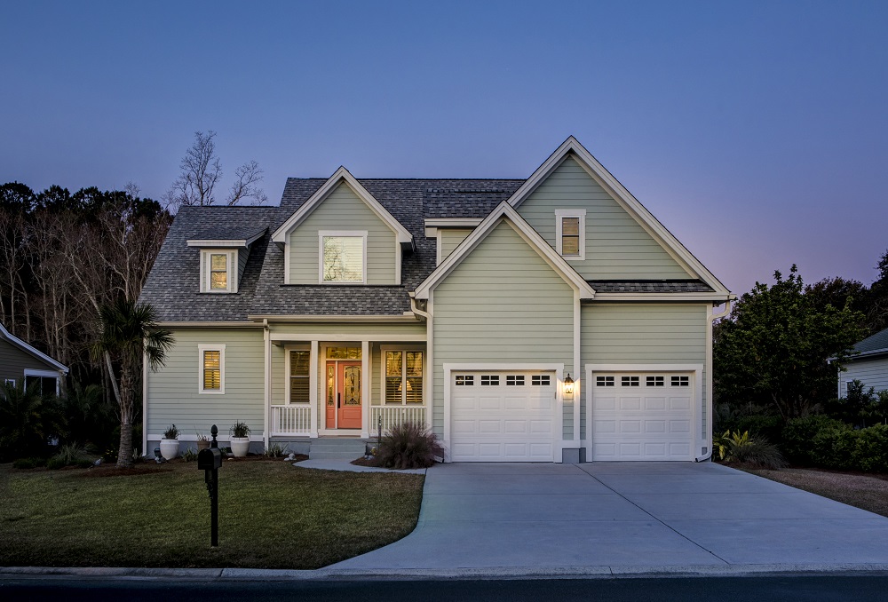 Two-story light green house with front porch, double garage featuring a recent garage door replacement Lakewood, and a well-maintained lawn at dusk.
