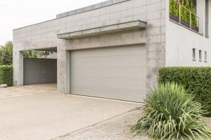 Modern house with light gray tiled exterior, a large closed garage door, a covered driveway, and landscaping with shrubs and ornamental grass—ideal for those considering garage door replacement in Lakewood.