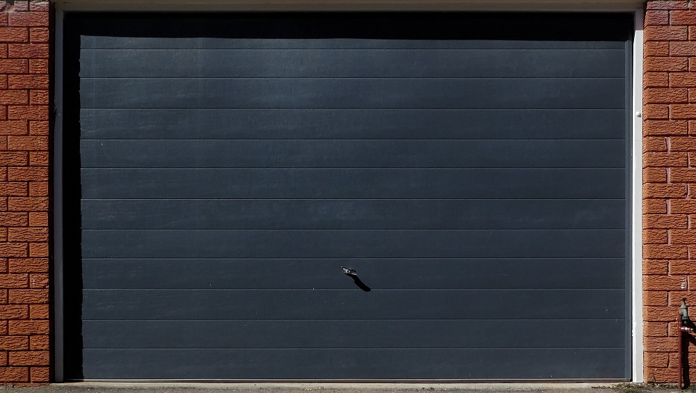 	
A closed black garage door with a small dent, likely from hailstorms in Parker, and a handle near the bottom, flanked by red brick walls.