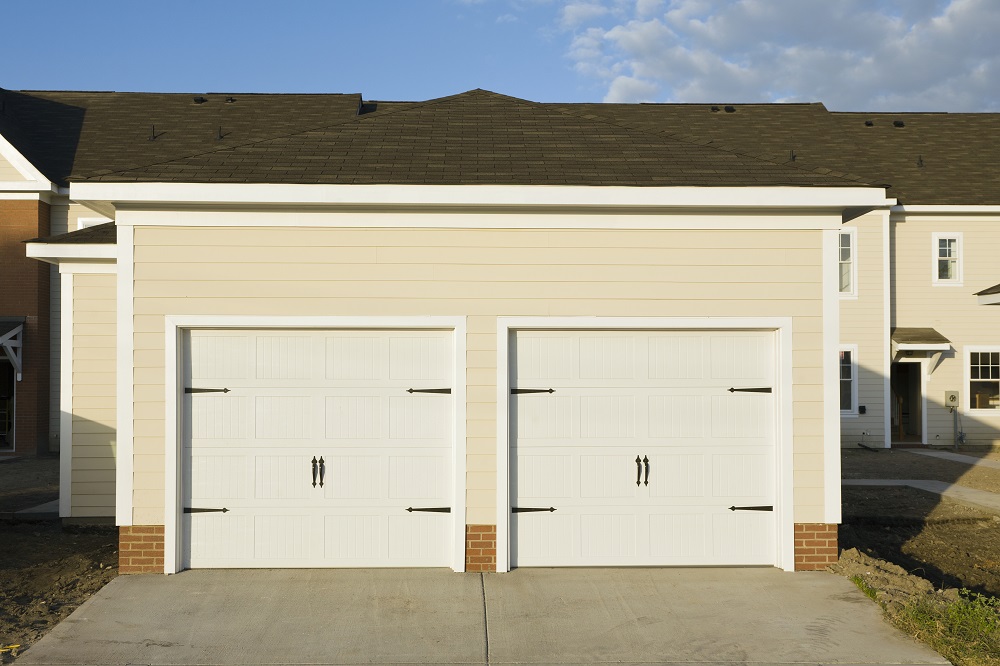A beige house with two white garage doors—one of the most popular garage door colors in Parker subdivisions—each featuring black metal handles and brick accents at the base, set on a concrete driveway.