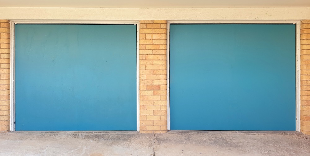 Two closed blue garage doors set in a yellow brick wall with a concrete driveway in front—an ideal example for those considering garage door replacement in Lakewood.