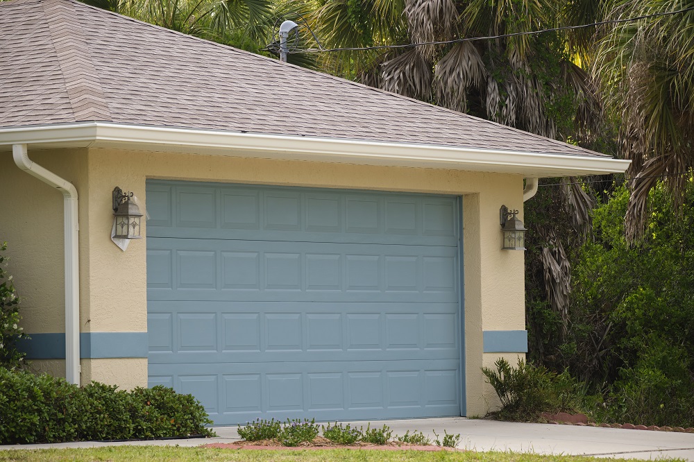  A beige house with a blue double garage door, two exterior wall lanterns, and surrounding greenery, designed to comply with HOA rules for garage doors in Lakewood.