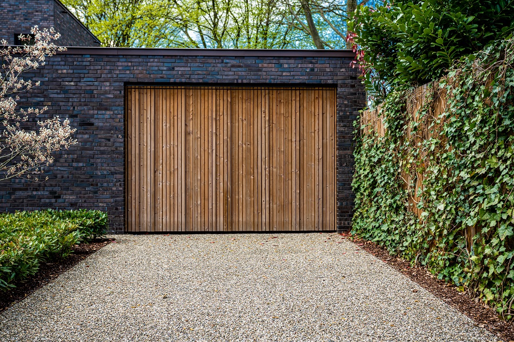 A modern garage with a wooden door, inspired by trending garage door styles in Greenwood, is surrounded by brick walls, green plants, and a gravel driveway.