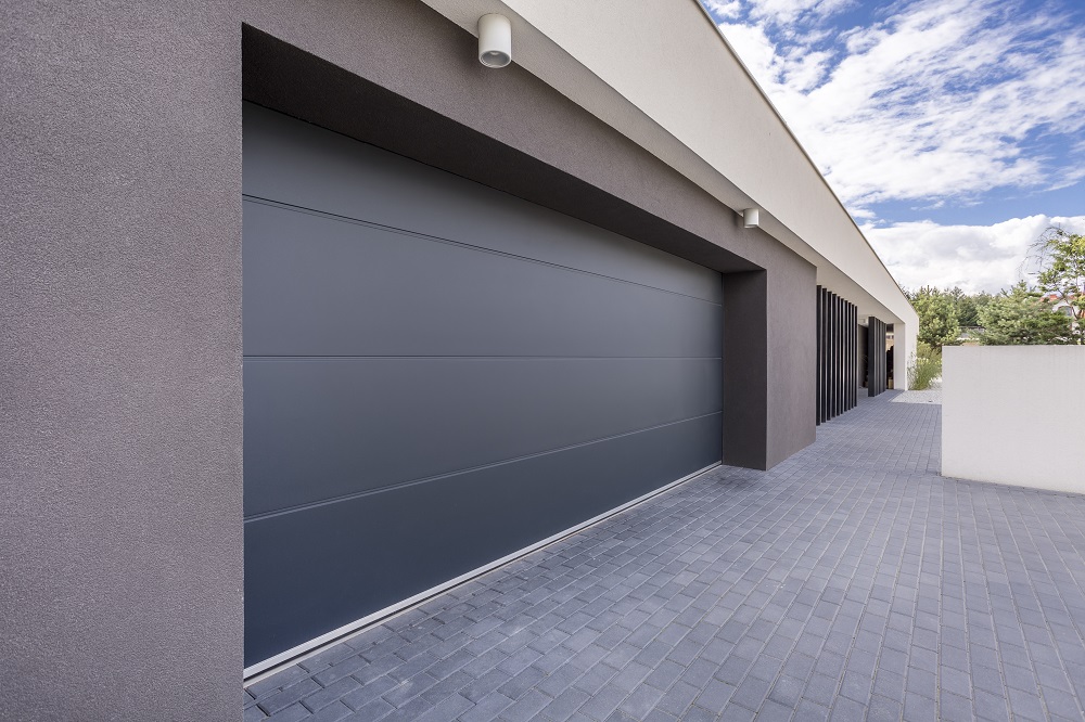 	
Modern house exterior with light gray stone walls, a large garage door reflecting Trending Garage Door Styles in Greenwood, covered driveway, green shrubs, and a paved driveway.
