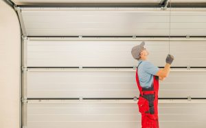 A worker in red overalls and a gray cap inspects or adjusts the cable of a raised garage door during a garage door installation in Greenwood.