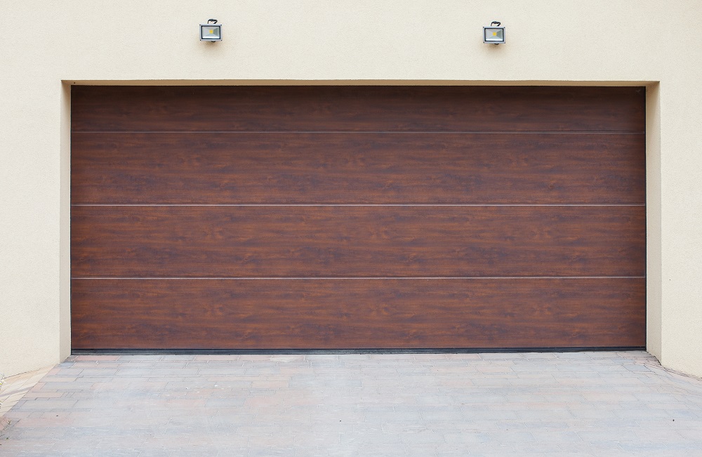 Closed brown wooden garage door set in a light beige wall, with two small outdoor lights above and a paved driveway—perfect inspiration for anyone considering Garage Door Replacement in Greenwood.