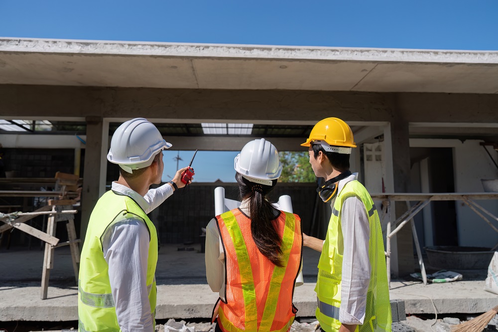 Three construction workers wearing safety vests and helmets stand at a building site, discussing garage door installation in Greenwood, as one person points toward the structure.