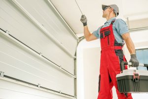A technician in red overalls and gloves repairs a garage door, holding a toolbox and adjusting a cable with a tool—showcasing expert Garage Door Replacement in Greenwood.