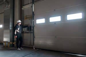 A person wearing a hard hat and work clothes presses a button to open a large industrial garage door with rectangular windows, typical of a slow opening garage door in Parker.