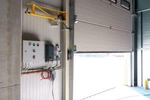 A partially open industrial garage door, resembling a slow opening garage door in Parker, stands next to a wall with control panels, wiring, and a mounted yellow safety bar inside a warehouse.
