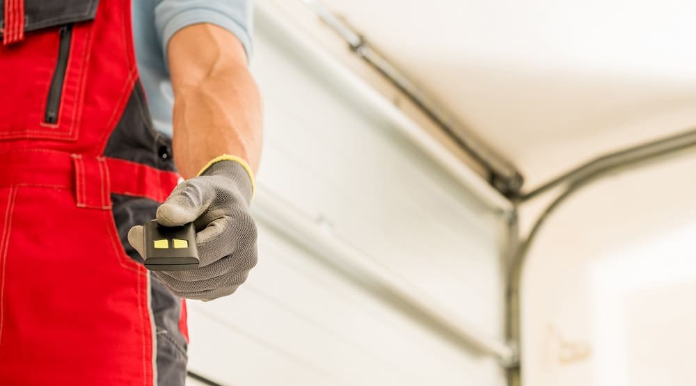A person wearing red overalls and gloves holds a Garage Door Opener Greenwood remote control in front of an open garage door.
