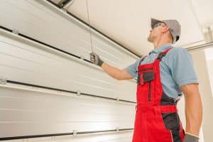 A worker in red overalls and a cap inspects or repairs a slow opening garage door in Parker using a metal tool, inside a well-lit garage.