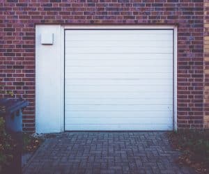 A closed white garage door, equipped with a Garage Door Opener Greenwood system, sits in a brick wall with a paved driveway in front and a small trash bin visible on the left side.