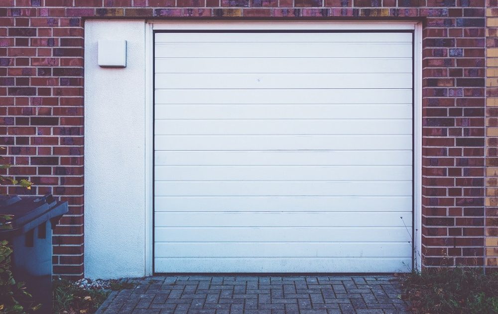 Closed white garage door set in a brick wall, with a paved driveway in front and a trash bin partially visible on the left—an example of the neat, reliable garage doors Lakewood homeowners appreciate.