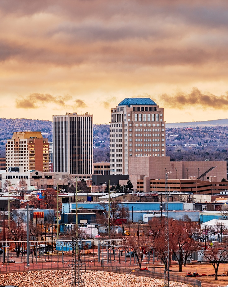 Downtown cityscape with several tall office buildings, leafless trees, and mountains in the background under a cloudy sky at sunset.