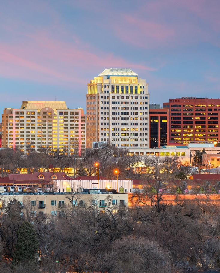 Downtown cityscape at sunset with illuminated office buildings, leafless trees in the foreground, and a sky with pink clouds—perfectly reflecting the growth in Commercial Garage Door Installation in Greenwood Village.
