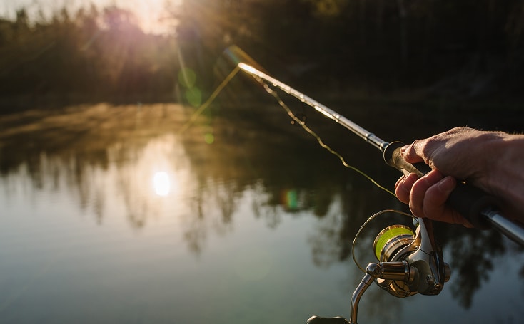 A person’s hand holding a fishing rod over a calm lake at sunset, with trees and sunlight reflecting on the water—much like the peaceful assurance that comes after a successful Commercial Garage Door Installation in Greenwood Village.