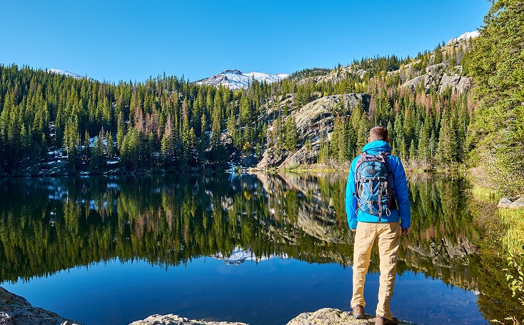 A person with a backpack stands on a rock overlooking a calm mountain lake in Lakewood, CO, surrounded by pine trees, with blue sky and snow-capped peaks in the background.