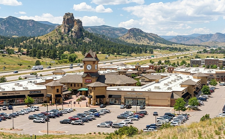 Shopping center with a clock tower and full parking lot, set against a backdrop of mountains and a highway under a partly cloudy sky.