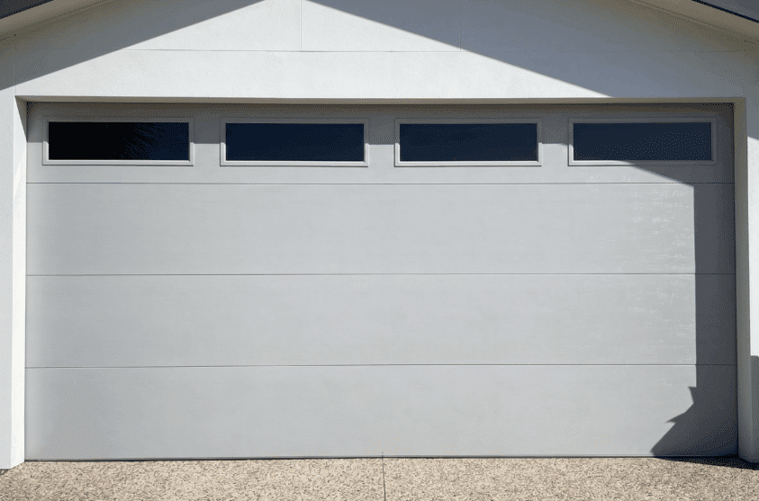 A closed modern garage door with four rectangular windows at the top, set in a white exterior wall above a pebble-textured driveway.