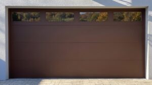 Closed brown garage door with four rectangular windows at the top, set in a white exterior wall with a paved driveway in front.