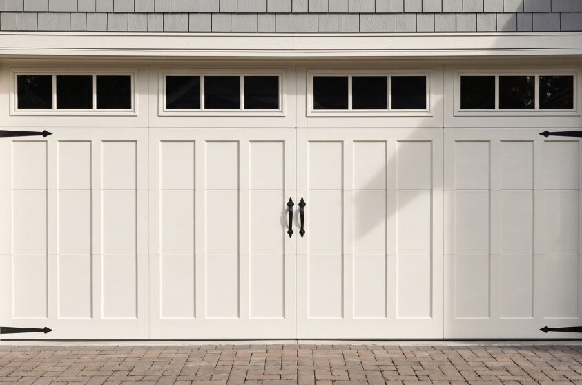 Closed white garage door with vertical panels, black decorative hinges, handles, and four rectangular windows along the top. Brick driveway and partial view of house siding and stone trim.