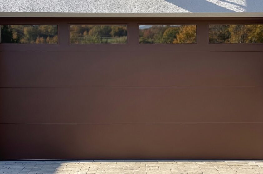 Closed brown garage door with four rectangular windows at the top, set in a white exterior wall with a paved driveway in front.
