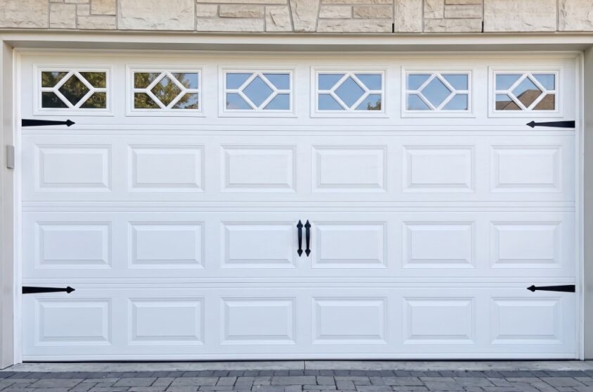 White garage door with four decorative windows at the top, black handles, and black hinge accents, set in a stone exterior wall.