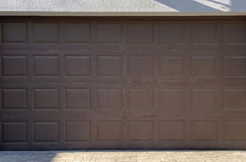 A closed brown sectional garage door with a rectangular panel design, set in a light-colored exterior wall above a paved driveway.