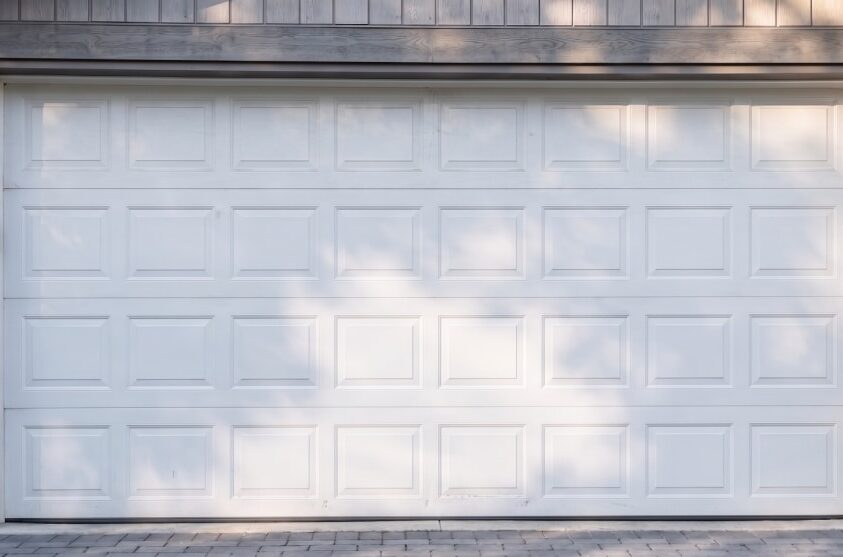 Closed white residential garage door with rectangular panels, framed by light stone and wood trim, with faint shadows visible on the surface.