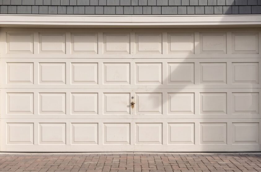 Closed beige garage door with rectangular panels, surrounded by grey siding and stone trim, set above a brick driveway.