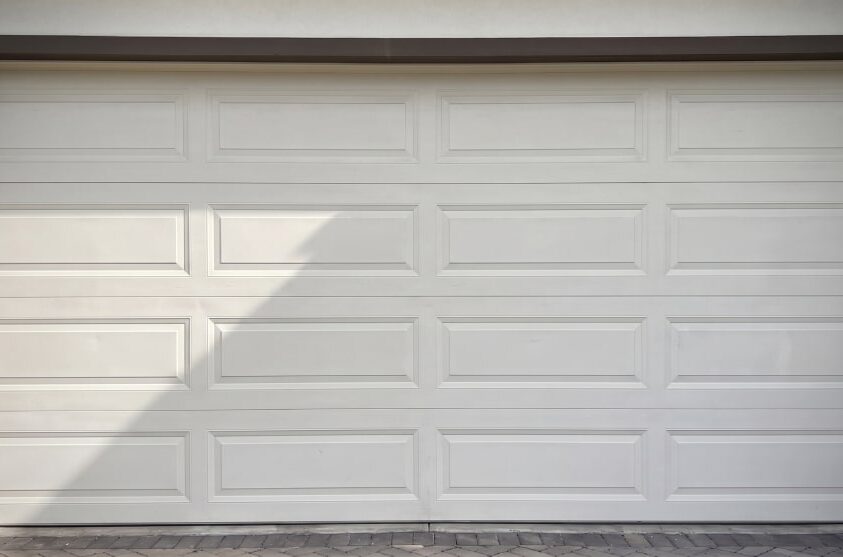 Closed white sectional garage door with rectangular panel design, set in a beige frame above a paved driveway.