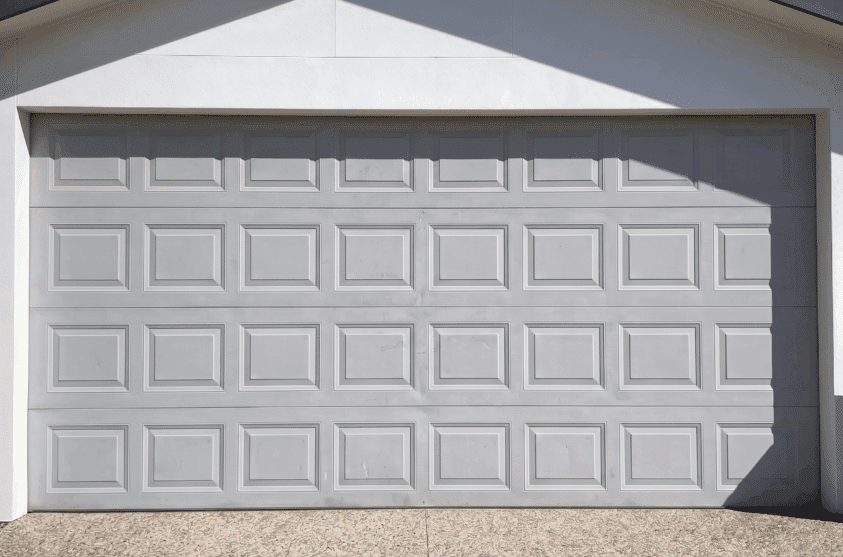 A closed, light gray sectional garage door with rectangular panels, set in a white exterior wall and illuminated by sunlight.