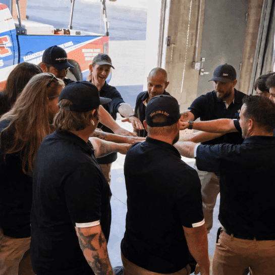 A group of workers in uniforms stand in a circle, each with one hand extended to the center in a gesture of teamwork inside an industrial or warehouse setting.