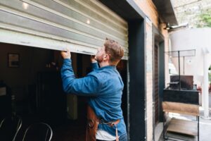 A man in a denim shirt and apron lifts a metal garage-style door to open a storefront or workshop.