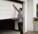 A man in a plaid shirt manually opens or closes a white garage door attached to a white building.